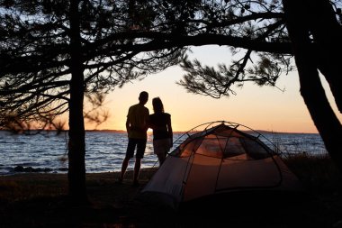 Genç turist çift erkek ve kadın ayakta mavi küçük çadır önünde göl kıyısında kucakladı karanlık siluetleri Gölü su arka plan zevk sessiz akşam temizleyin. Turizm ve kamp kavramı.