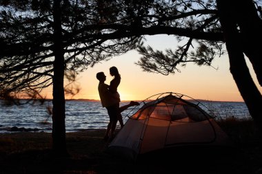 Genç turist çift erkek ve mavi küçük çadır önünde deniz kıyısında kucakladı duran kadın siluetleri deniz su arka plan zevk sessiz akşam temizleyin. Turizm ve kamp kavramı.