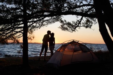 Çadır mavi berrak su arka planda sessiz akşam zevk yakınındaki deniz kıyısında siluetleri genç turist çift erkek ve kadın öpüşme ve ayakta kucakladı. Turizm ve kamp kavramı.