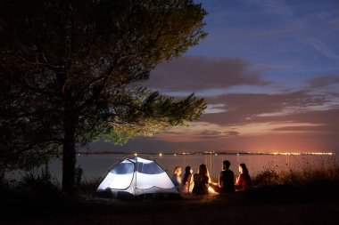 Gece yaz göl kıyısında kamp. Beş genç turist beach yakınındaki güzel mavi akşam gökyüzü altında çadır kamp ateşi etrafında oturan grup. Turizm, dostluk ve doğa kavramının güzellik.