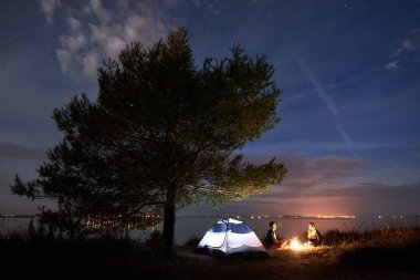 Küçük turistik çadır altında gölgeli büyük ağaç ve Genç çift, sakallı adam ve gece şenlik ateşi mavi akşam gökyüzü altında tarafından göl kıyısında oturan güzel kadın. Turizm, kamp ve sevgi dolu ilişkiler kavramı