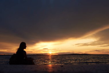 Su kenarında, sahil üzerinde tek başına oturmak sırt çantası ile profil turist kadının güzel bir günbatımı manzarası karanlık akşam gökyüzü arka plan üzerinde silüeti. Turizm ve tatil kavramı.