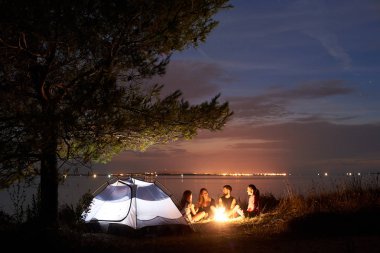 Grup dört turist turist çadır altında ağacında, önünde şenlik ateşi, deniz kıyısında parlak mavi akşam gökyüzü temiz su arka plan üzerinde işaret sakallı adam oturuyor. Turizm ve kamp kavramı.