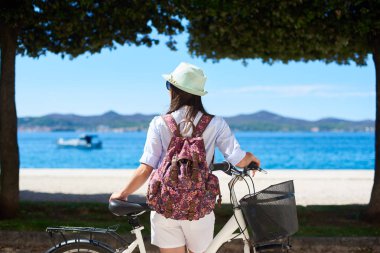 Kapanış, sunhat beyaz kadın turist görünümü geri ve vizyon ağaçlar ve dağlar karşı kıyıya arka plan üzerinde altında kıyısından kaldırımda Bisiklet ile ayakta sırt çantası. Turizm ve tatil kavramı.