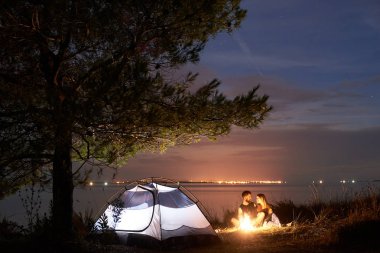 Gece sahil kamp. Genç turist çift, sakallı adam ve deniz kıyısında kamp ateşi Turizm çadır büyük ağacın altında yanan yakın oturan güzel kadın. Turizm, kamp ve sevgi dolu ilişkiler kavramı.