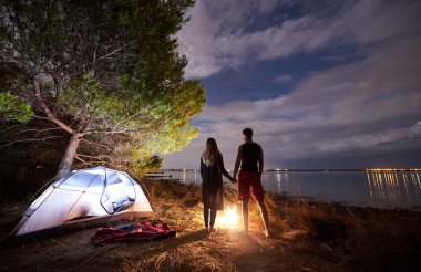 Arkadan görünüşü genç turist çift, atletik bir adam ve gökyüzü ve deniz su arka plan akşam tutuşup turist çadır yakınındaki ateşin yanında duran ince kadın. Aktif yaşam tarzı ve sevgi dolu ilişkiler kavramı.