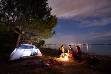 Grup üç turist, şenlik ateşi yakın turist çadır ağacın altında göl kıyısında oturan kadın ve genç adam. Sessiz su yüzeyi ve akşam gökyüzü arka plan üzerinde. Turizm, dostluk ve kamp kavramı.