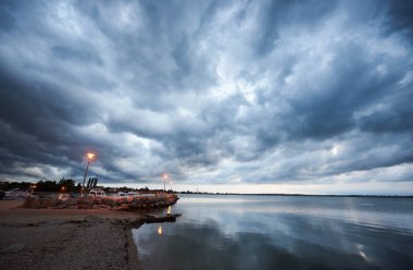 Güzel manzara sahil resort Town alacakaranlıkta. Cruise motorlu tekne kıyıda bağlantılı yüzen, bulutlu gökyüzü ve sokak lambaları sakin mavi denizde yansıtan parlayan su, evler çatılar yeşil ağaçların arkasında.