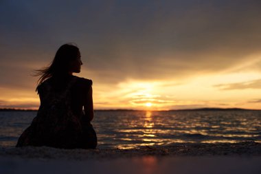 Sahil su kenarında, güzel bir günbatımı manzarası karanlık akşam gökyüzü arka plan üzerinde zevk üzerinde tek başına oturmak sırt çantası ile turist kadın silüeti görünümü geri. Turizm ve tatil kavramı.