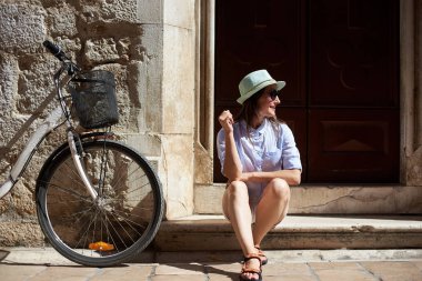 Güneş gözlüğü, beyaz şort, bluz ve bisiklet binasında eski antik parlak güneşli gün yüksek kaldırım üzerinde oturan şapka çok gülümseyen uzun saçlı kız. Gezi ve aktif yaşam tarzı kavramı.