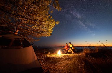 Mavi yıldızlı gökyüzü altında gölde kamp gece çadır. Genç turistler, erkek ve kadın üzerinde kamp ateşi ısınma uzak kıyı ufuk arka plan su ve şehir ışıkları temizleyin. Turizm ve sevgi kavramı.