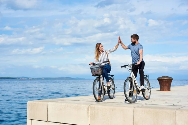Tourist couple, man and woman standing with bicycles on high paved stone sidewalk, giving high five each other on bright sunny day. Clear blue sea water on background. Active holiday concept.