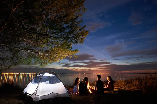 Group of four tourists sitting on sea shore at bonfire in front of tourist tent under tree, bearded man pointing at bright blue evening sky on clear water background. Tourism and camping concept.