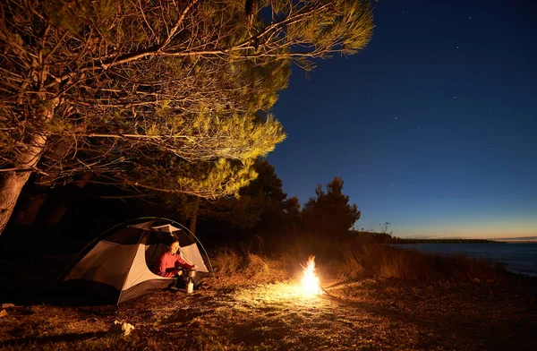 Night camping at seaside. Smiling tourist woman sitting in entrance of tent, preparing food on a gas burner, near campfire, enjoying beautiful view of starry sky. Active lifestyle concept.