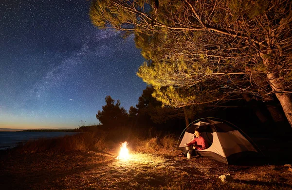 湖でのキャンプの夜 天の川の景色の下でガス バーナーの上に食べ物を準備する女性観光 キャンプファイヤーで大きな木の近くのテントの入り口で一休みを幸せな澄んだ青い水 — ストック写真