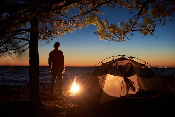 湖の海岸の下でのキャンプの夜は澄んだ青い空です 近く澄んだ水にテント 遠い地平線背景点灯キャンプファイヤーに単独で立っている観光女性の背面します レクリエーション — ストック写真