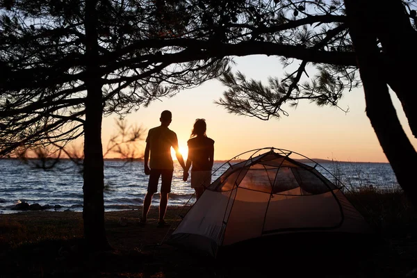 年轻的游客夫妇的黑暗剪影站在湖岸边的男人和女人牵着手在小帐篷前的明亮的黄色夕阳和水晶蓝色清澈湖水背景 — 图库照片