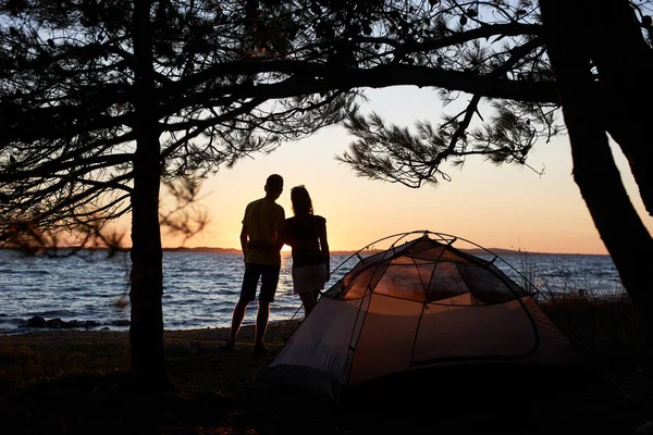 Dark silhouettes of young tourist couple man and woman standing embraced on lake shore in front of small tent on blue clear lake water background enjoying quiet evening. Tourism and camping concept.