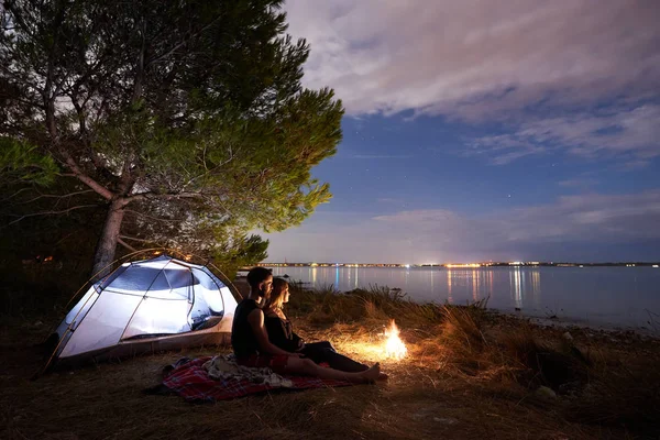 Small tourist tent and young couple, bearded man and pretty woman sitting embraced at night by fire watching quiet lake under blue evening starry sky. Tourism, camping and loving relations concept.