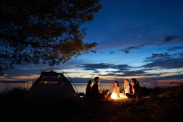 晚上夏天在湖岸边露营 一群五年轻的游客坐在海滩周围的篝火附近的帐篷在美丽的蓝色傍晚的天空下 友谊与自然之美 — 图库照片