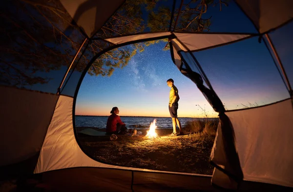 見る夕日観光テントの中 若い観光客カップル 男と女 たき火で休んで夜の星空や結晶海水背景に海海岸にガスバーナーの上に食べ物を準備します — ストック写真