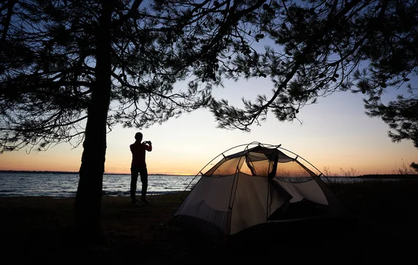 フォト カメラで観光テント前に海海岸に立って 夕暮れ時の静かな夏の夜澄んだ海の写真を撮る若いハイカー女性のシルエット キャンプのコンセプト — ストック写真