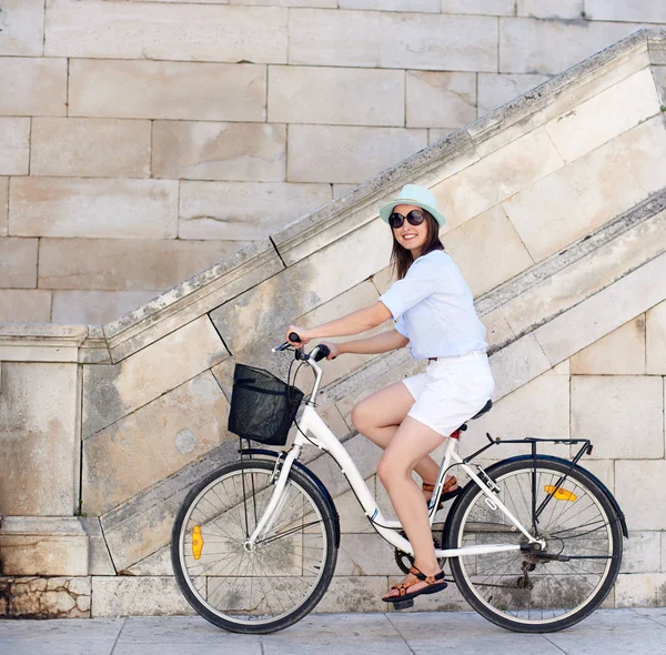 Happy woman in sunglasses, white shorts, blouse and hat riding on bicycle on white stone wall and stairs background on bright sunny day. Sightseeing and vacations concept.