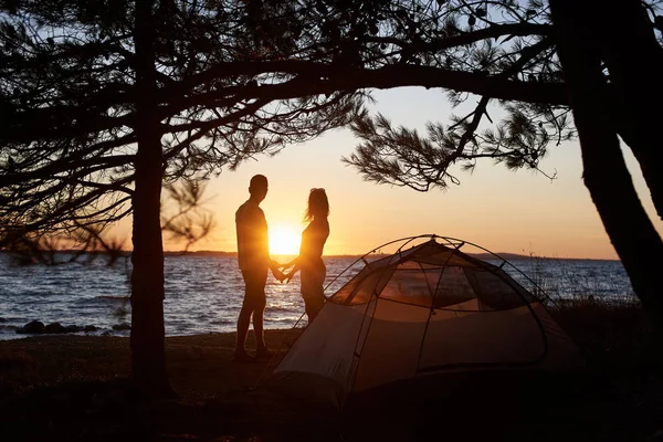 若い観光客カップル男と明るい黄色の太陽と結晶の明確な湖の青い水背景設定の小さいテントの前手を取り合って海海岸に立って女の暗いシルエット — ストック写真