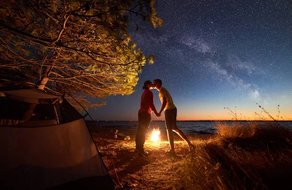 Night camping at sea under blue starry sky. Young couple, man and woman kissing at campfire. Clear water and city lights on distant shore on background. Tourism concept, outdoor activity concept