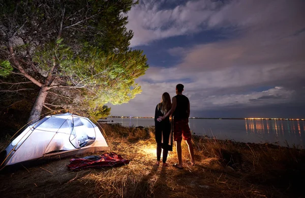 若い観光客カップル スポーツ人と受け入れ観光テントの火に照らされて夜の空と湖の水の背景に近く立っているスリムな女性の後ろ姿 アクティブなライフ スタイルと愛情のある関係の概念 — ストック写真