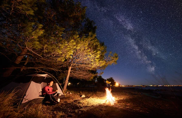 Night camping at seaside. Hiker woman preparing food on a gas burner, having a rest in entrance of tourist tent at campfire near forest against bright blue starry sky with Milky way