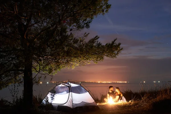 海辺の近くでキャンプの夜 若い観光客カップル 髭の男 大木の下で観光テントでキャンプファイヤーを燃焼の近くの海岸に座っているきれいな女性 キャンプと関係概念を愛する — ストック写真