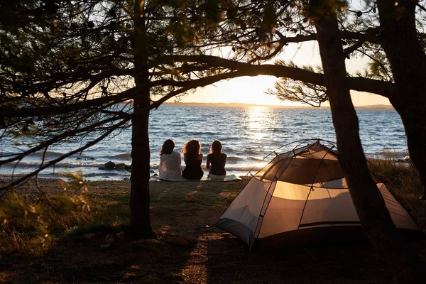 澄んだ水の背景に美しい夕日を楽しむ湖畔観光の木の下で テントの前に座っている 人の若い女性の背面します 友情およびキャンプのコンセプト — ストック写真