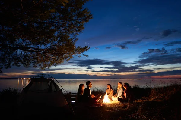 澄んだ水の背景に明るい青い夕空を指してアゴヒゲ観光の木の下で テントの前に焚き火で海岸に座って観光客を 人のグループ キャンプのコンセプト — ストック写真