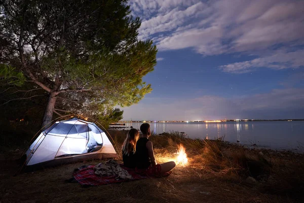 海岸のキャンプの夜 若いカップル ひげを生やした男性と曇り夜の青い空の下でのキャンプファイヤーで観光テント近く残りを持つきれいな女性 観光と愛情のある関係の概念 — ストック写真