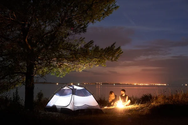 若いカップル 髭の男と夜に照らされて静かな水表面の背景に大きなツリーの下で観光テントでキャンプファイヤーを燃やして海海岸にキャンプで残りを有する女性ハイカー 観光コンセプト — ストック写真