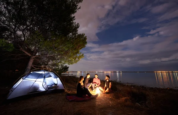 Group of four young tourists, men and women having a rest on sea shore at campfire near tourist tent. Quiet water surface and cloudy evening sky background. Tourism, friendship and camping concept.