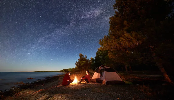 Night camping on shore. Young man and woman tourists sitting relaxed in front of tent at campfire under starry sky with Milky way on blue water and forest background. Active outdoor lifestyle concept