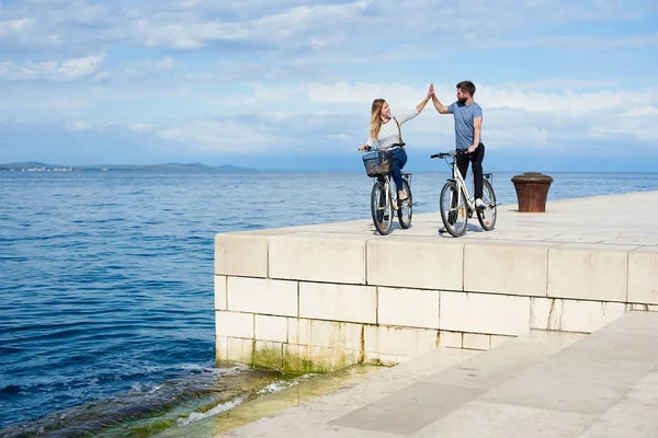 Tourist pair, man and woman standing with bicycles on high paved stone sidewalk, giving high five each other on bright sunny day. Clear blue sea water on background. Active holiday concept.