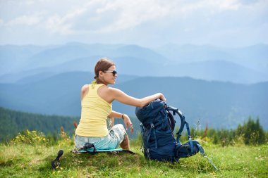Arkadan görünüşü ile sırt çantası, çimenli tepe üzerinde oturan çekici kadın turist güneş gözlüğü, Karpat Dağları yaz bulutlu gün zevk takıyor. Açık hava etkinliği, turizm kavramı