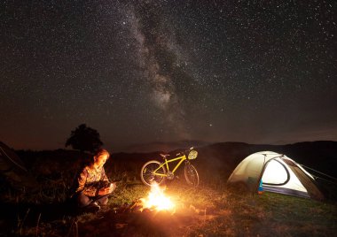Genç kadın turist kamp ateşi, ışıklı turist çadır, dağ bisikleti inanılmaz güzel akşam gökyüzünde yıldızlar ve Samanyolu dolu altında yanan yakınındaki gece kamp dinlenme. Turizm kavramı