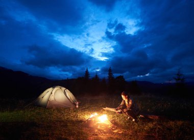 Genç kadın turist dağda kamp, kamp ateşi ve ışıklı turist çadır güzel bir akşam bulutlu gökyüzü altında yanan yakın oturan gece zevk. Turizm, açık hava etkinliği konsepti