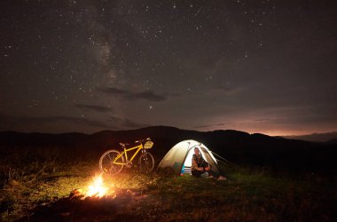 Genç kadın yolcu gece kamp ateşi, ışıklı turist çadır, dağ bisikleti yıldızlarla dolu güzel bir akşam gökyüzü altında yanan yakınındaki kampa adlı bir dinlenme. Açık kavramı, etkinlik ve Turizm
