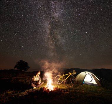 Genç kadın bisikletçi gece kamp kamp ateşi, ışıklı turist çadır, dağ bisiklet inanılmaz güzel akşam gökyüzünde yıldızlar ve Samanyolu dolu altında yanan yakınındaki zevk. Astrophotography