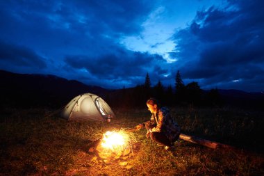 Kamp ateşi ve ışıklı turist çadır güzel bir akşam bulutlu gökyüzü altında yanan yakın oturan dağlarda gece kamp, bir dinlenme aktif kadın yolcu. Turizm, açık hava etkinliği konsepti