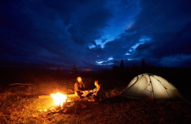 Kamp ateşi ve ışıklı turist çadır güzel bir akşam bulutlu gökyüzü altında yanan yakın oturan dağlarda gece kamp, bir dinlenme mutlu çift erkek ve kadın gezginler. Turizm kavramı