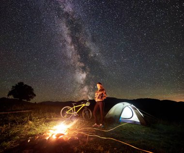 Genç kadın bisikletçi gece kamp ateşi, ışıklı turist çadır, dağ bisiklet inanılmaz güzel akşam gökyüzünde yıldızlar ve Samanyolu dolu altında yanan yakınındaki kampa adlı bir dinlenme. Astrophotography