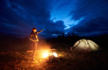 Bulutlu gökyüzü akşam itibariyle işaret eden genç bir kadın yolcu kamp ateşi ve ışıklı turist çadır, yanan yakın duran dağlarda gece kamp dinlenme. Turizm, açık hava etkinliği konsepti