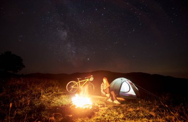 Genç kadın yolcu gece kamp ateşi, ışıklı turist çadır, dağ bisikleti yıldızlarla dolu güzel bir akşam gökyüzü altında yanan yakınındaki kampa adlı bir dinlenme. Açık kavramı, etkinlik ve Turizm