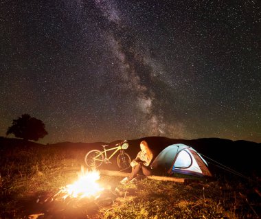 Genç kadın turist gece kamp ateşi, ışıklı turist çadır, dağ bisiklet inanılmaz güzel akşam gökyüzünde yıldızlar ve Samanyolu dolu altında yanan yakınındaki kampa adlı bir dinlenme. Astrophotography